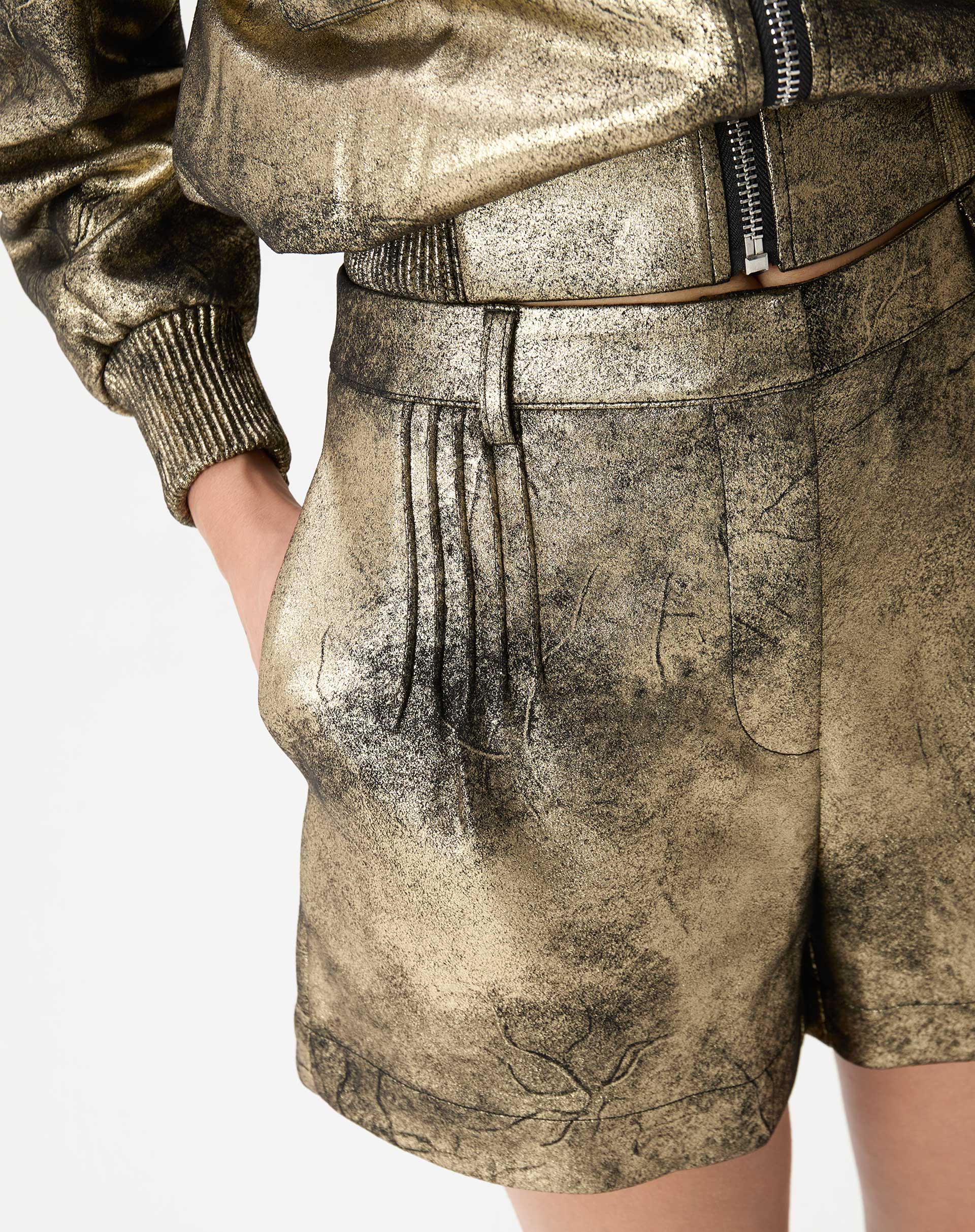 Close nas pregas frontais e bolso lateral do shorts metálico dourado com pespontos visíveis