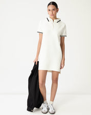 Vestido polo tricot off-white curto em modelo frontal segurando jaqueta preta, gola polo com filete preto e manga curta — look casual minimalista